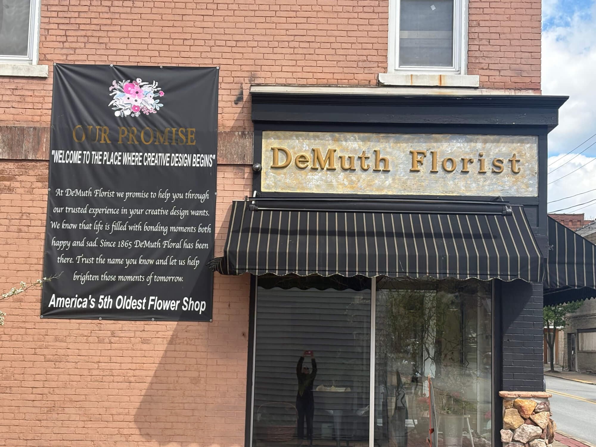 Exterior of DeMuth Florist shop displaying a banner with the slogan "Welcome to the place where creative design begins" and noting it as America's 5th oldest flower shop.