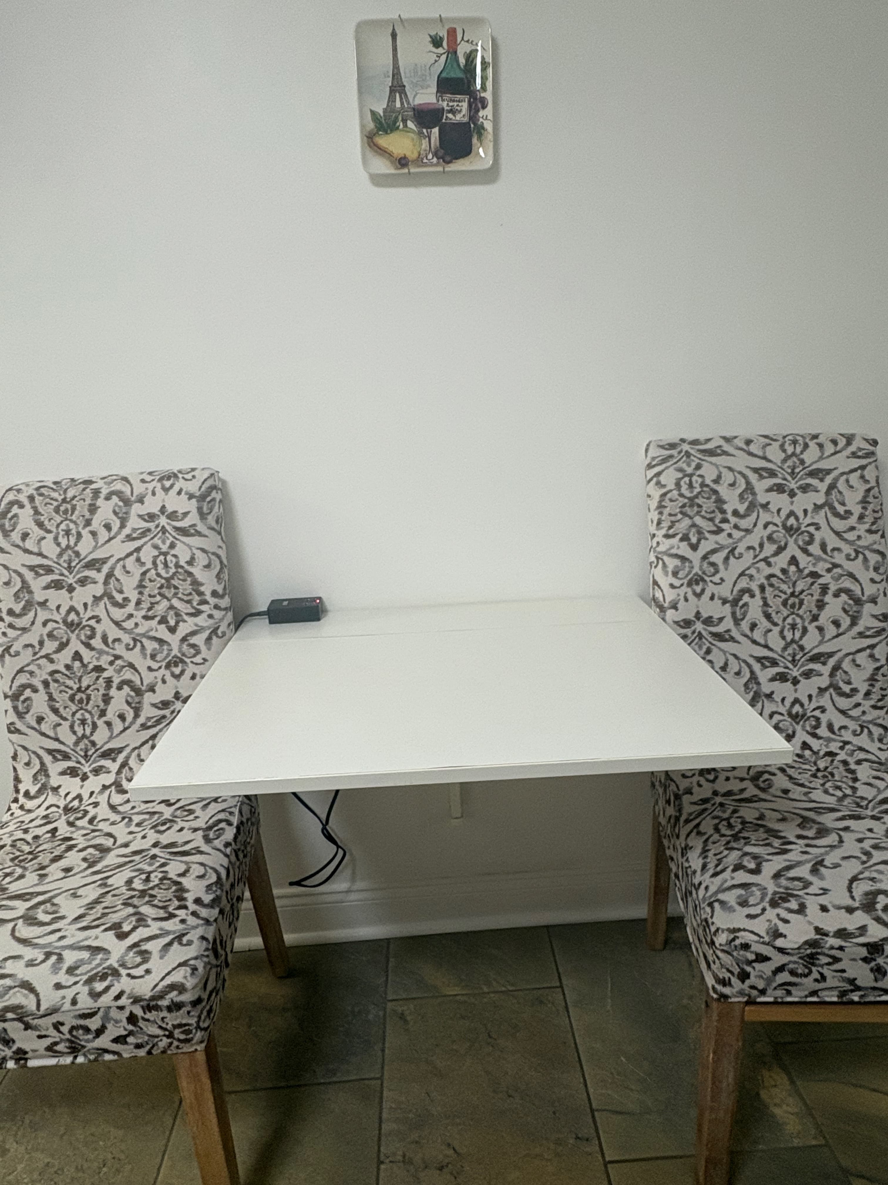 Drop down table is between two beige wi th black printing on cushioned chairs. USB port is on table. Above table is porcelain plate with images of Eiffel Tower, wine bottle, wine glass filled with wine and cheese