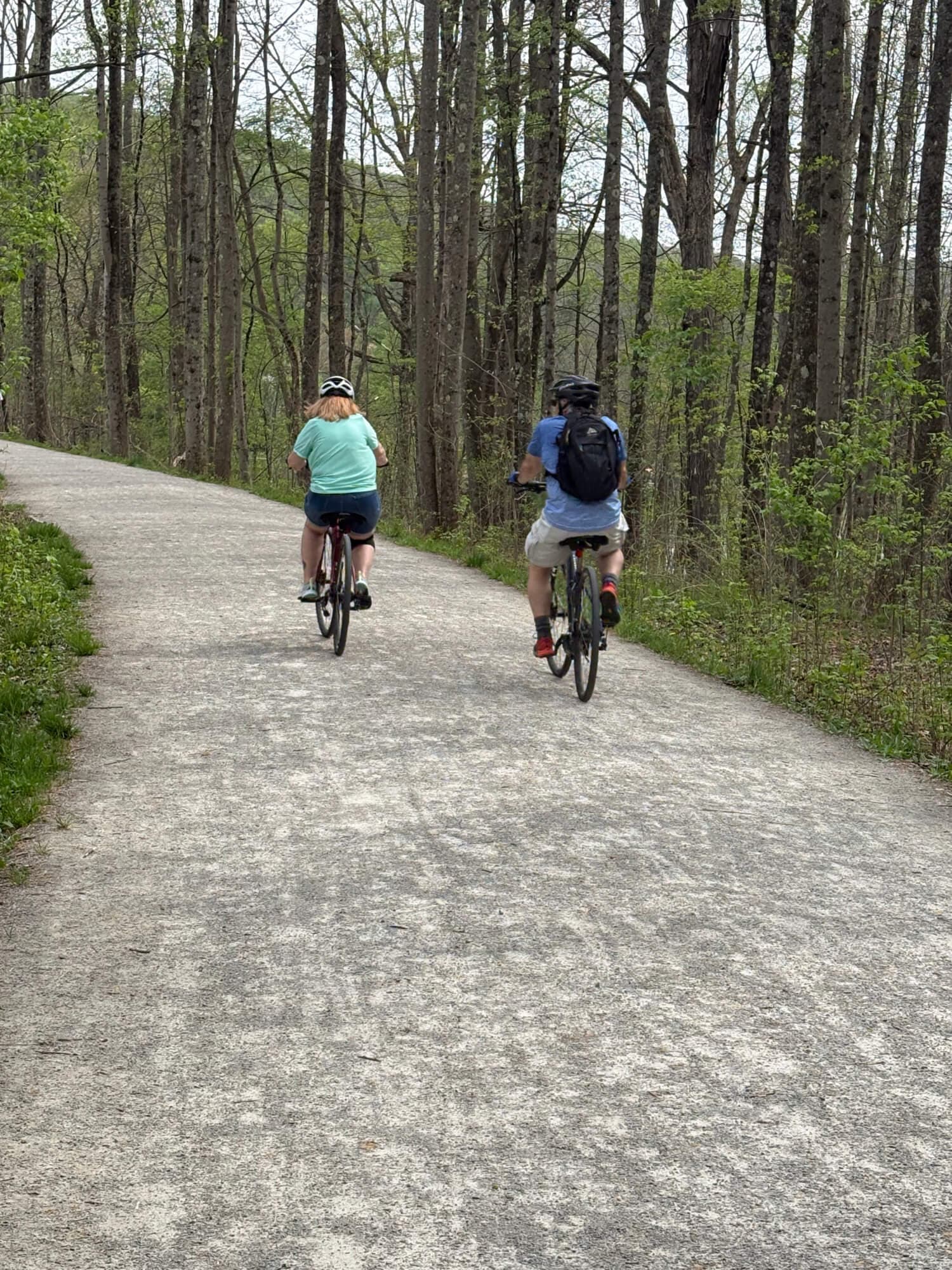 Two cyclists ride along a dirt path in a wooded area.