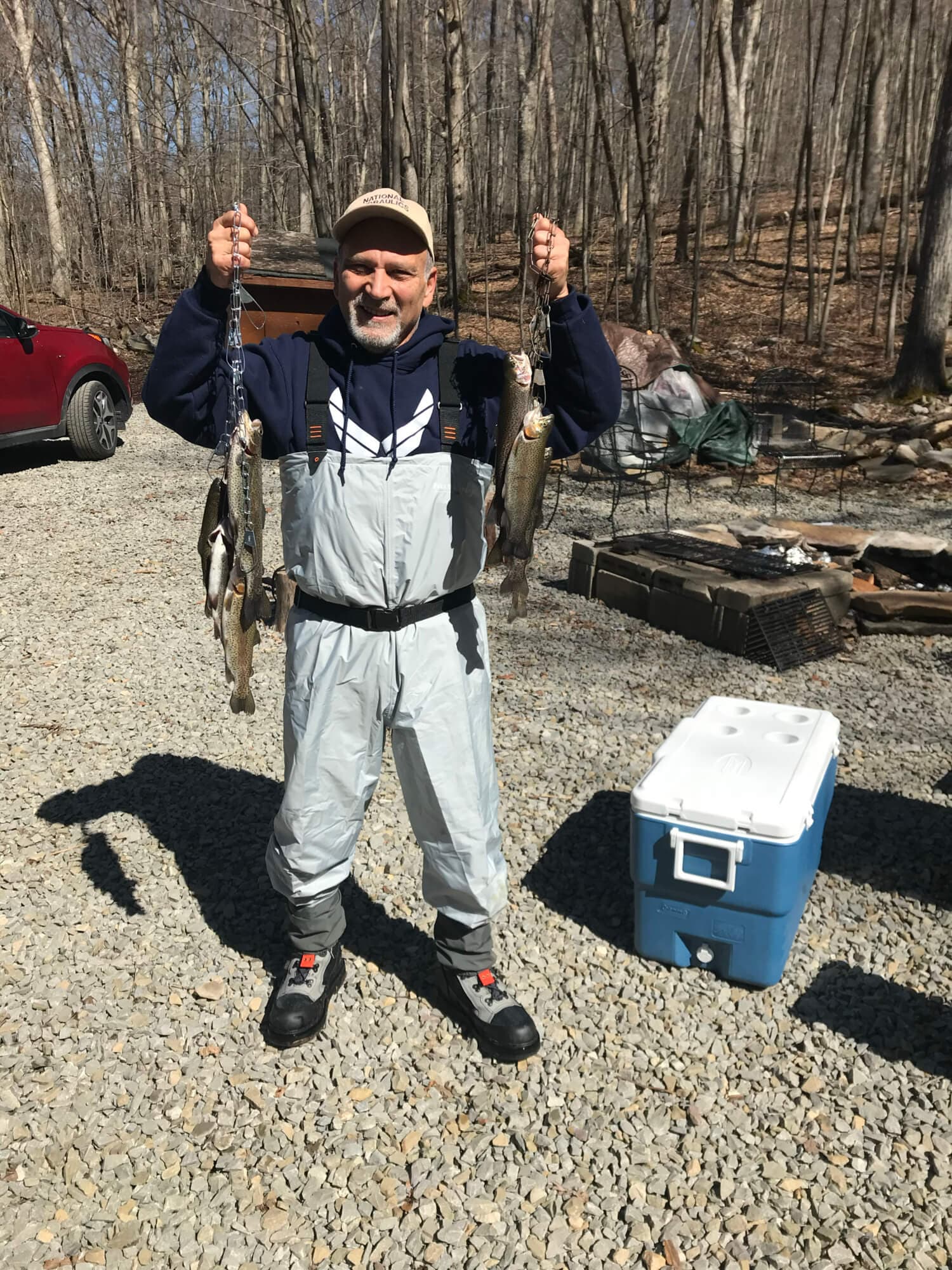 A man in waders holds up several fish while standing on gravel near a cooler.
