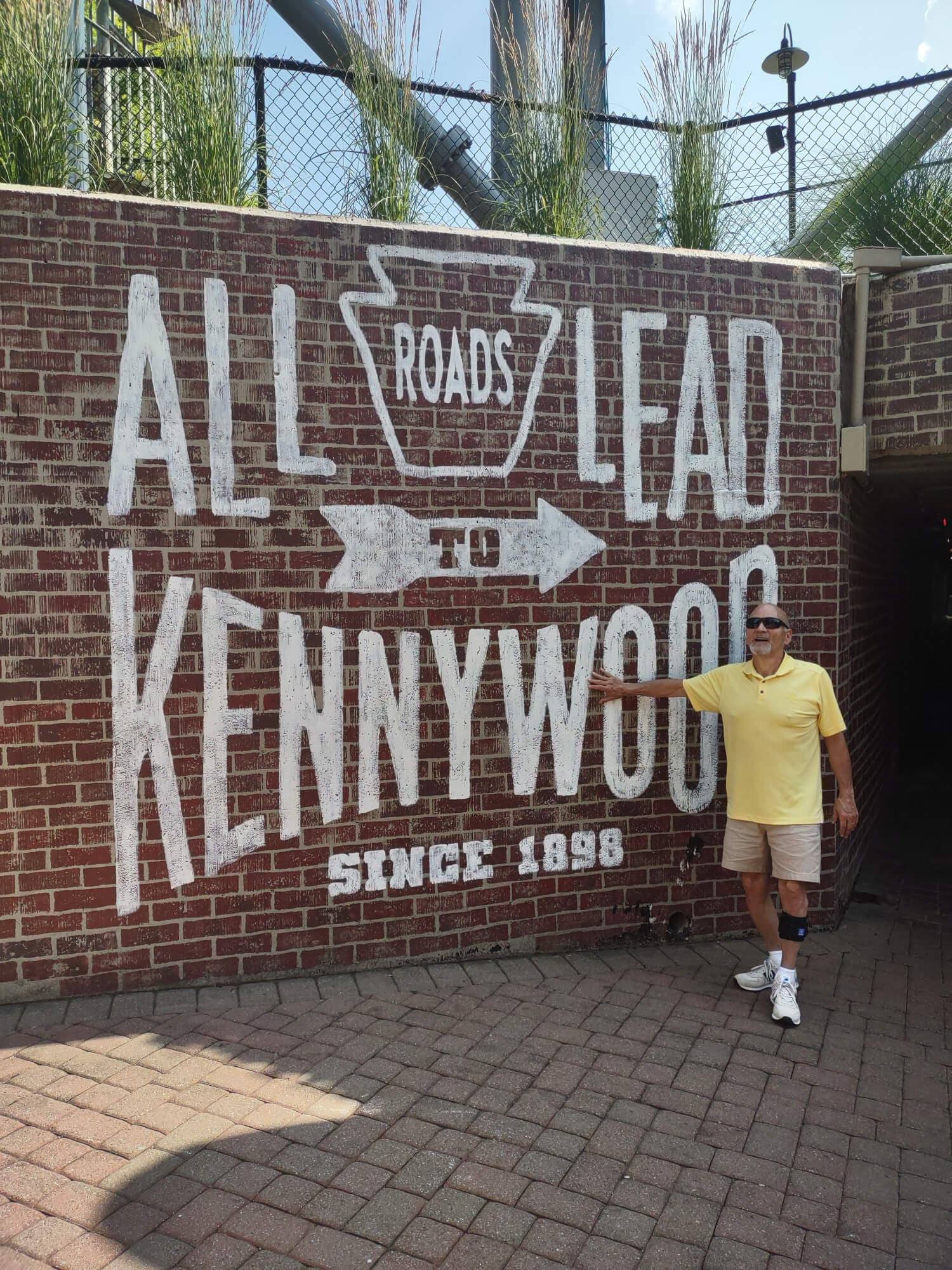 A man in a yellow shirt stands beside a brick wall featuring the sign "All Roads Lead to Kennywood Since 1898."