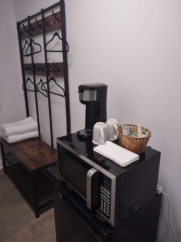 Coffee maker with cups and and basket of Keurig cups, assorted teas on top of black microwave on black mini-refrigerator. Dark wood and iron coat stand has 6 clothes hangers, shell with 2 white blankets.