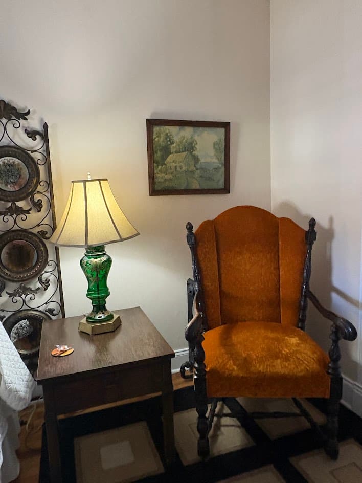 By the bed sits a green lamp with gold design shade is ivory shade. Jacobean chair walnut wood lathed, antique picture in back of chair