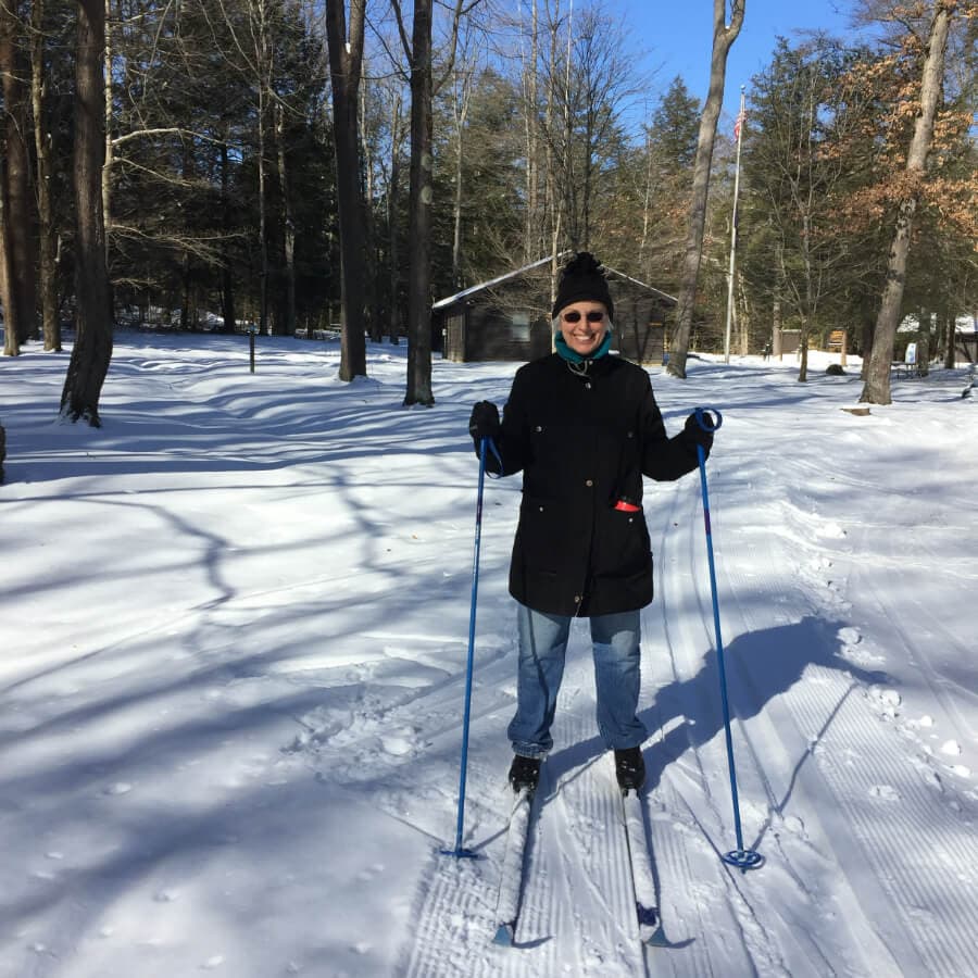 A person stands on ski poles in a snowy forested area, wearing a black coat and a hat.