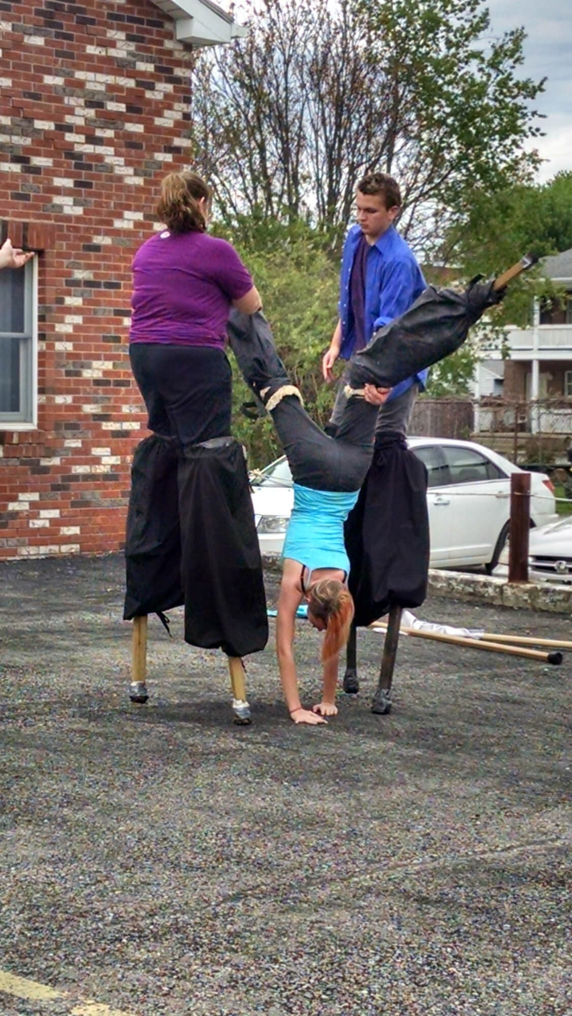Two people assist a third, who is performing a handstand on a pair of tall, black-covered structures in an outdoor setting.