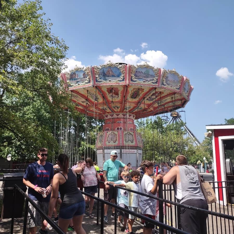A busy amusement park ride with a colorful swing carousel and groups of people waiting in line.