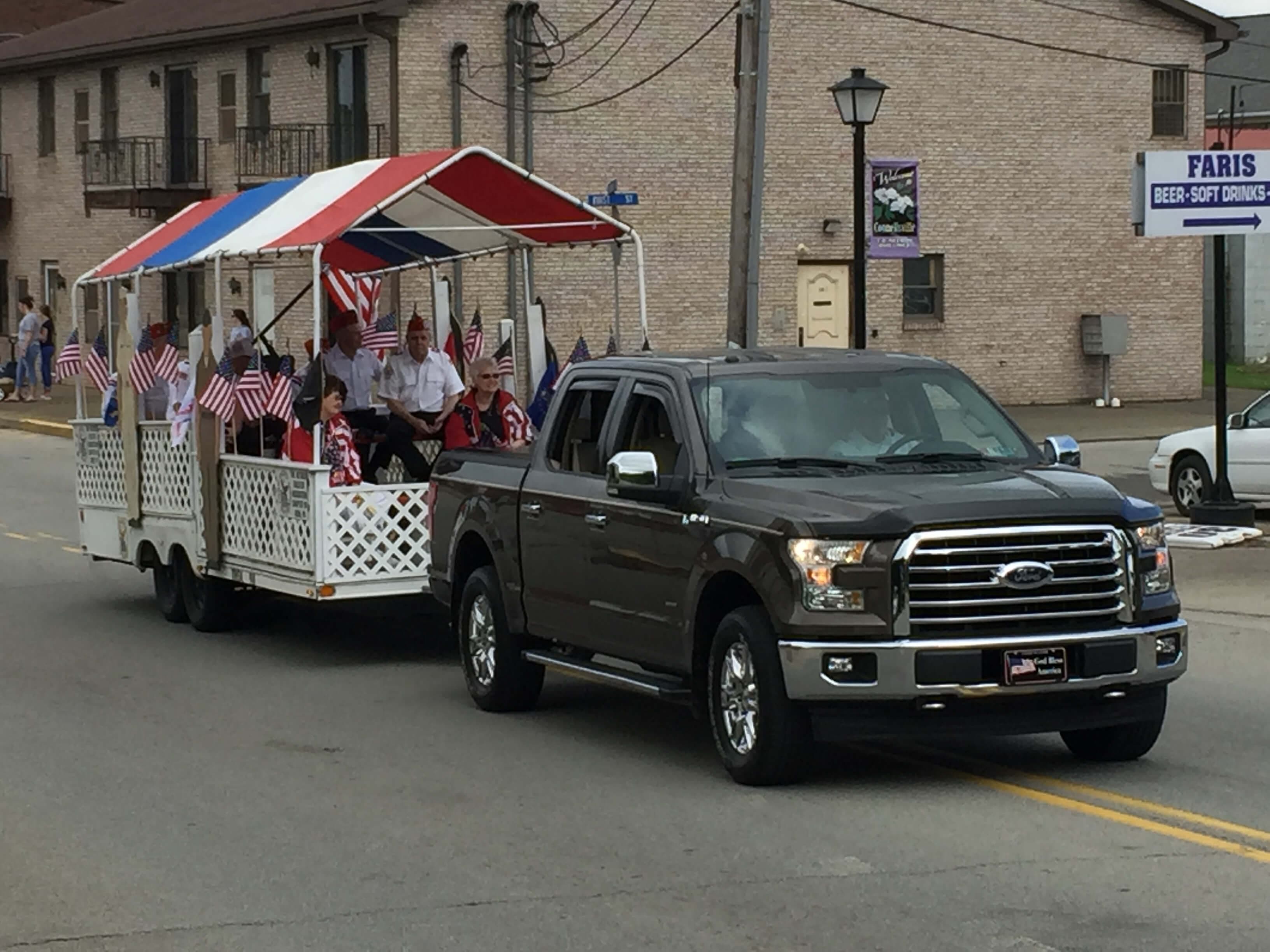 A gray pickup truck tows a decorated trailer with people celebrating while waving small American flags in a parade setting.